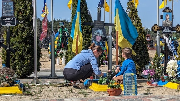 Junge Frau und kleiner Junge knien zwischen ukrainischen Flaggen am Grab des gefallenen Familienvaters