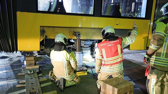 Feuerwehrleute stehen und knieen vor einer Straßenbahn. Eine hydraulischer Kolben hebt eine Bahn an. 