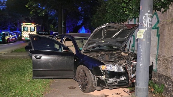 Ein Auto steht demoliert mit offener Motorhaube an einem Mast, im Hintergrund sieht man einen Krankenwagen und einen Streifenwagen.