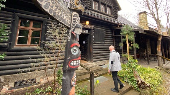 Holzhaus mit Aufschrift "Villa Bärenfett", davor Marterpfahl und Besucher