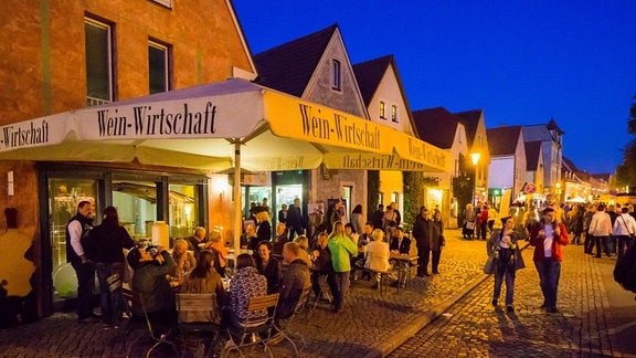 Menschen sitzen und gehen spazieren vor einem Weinlokal