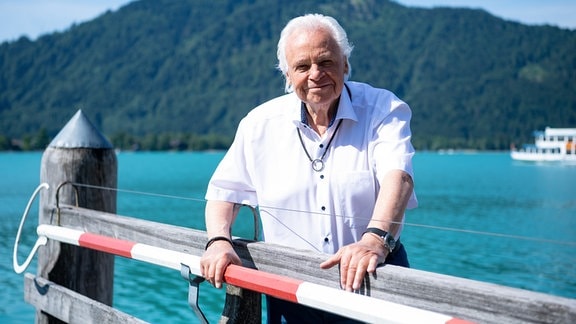 Ein älterer Herr mit weißem Haar steht an einem Geländer, im Hintergrund das blaue Wasser eines Sees, dahinter ein bewaldeter Berg