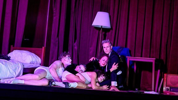 Vier teilweise leicht bekleidete Schauspieler schmiegen sich liegend an einen sitzenden Schauspieler. Das Bühnenbild zeigt ein Schlafzimmer.