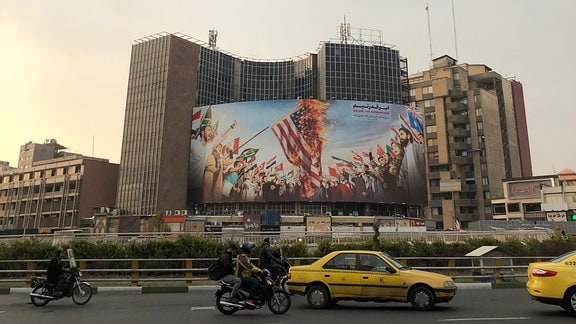 Eine vielbefahrene Straße in Teheran, im Hintergrund ein Plakat auf dem die us-amerikanische Flagge verbrannt wird.