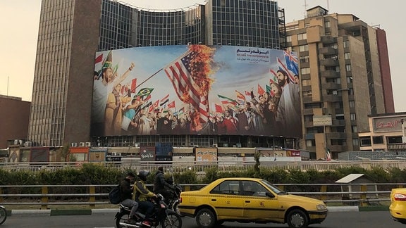 Eine vielbefahrene Straße in Teheran, im Hintergrund ein Plakat auf dem die us-amerikanische Flagge verbrannt wird.