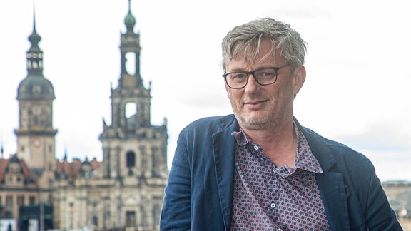Matthias Pfitzner, ein Mann mittleren Alters mit grauen Haaren, Dretagebart und Brille, trägt ein gemustertes Hemd mit blauem Sakko. Im Hintergrund ist die Stadtsilhoutte von Dresden zu sehen.
