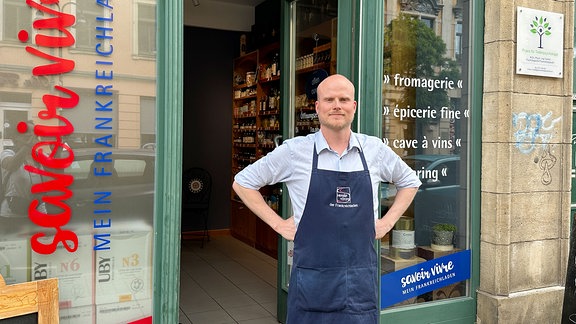 Ein Mann mit blauer Schürze steht vor seinem Ladengeschäft.