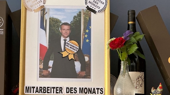 Mitarbeiter des Monats steht auf einem Bilderrahmen in dem ein Bild des französichen Präsidenten Macron zu sehen ist.