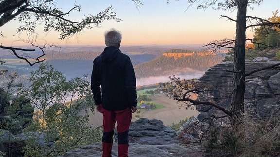 Tom Pauls von hinten steht auf einem Berg und schaut über eine weite Landschaft.