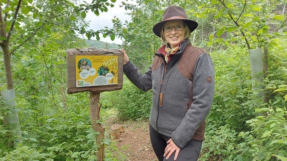 Eine Frau mit Rangerhut steht im Wald an einem Schild und blickt lächelnd in die Kamera. Es ist die Unternehmerin und Waldbesitzerin Bärbel Kemper in Liebstadt in der Sächsischen Schweiz.