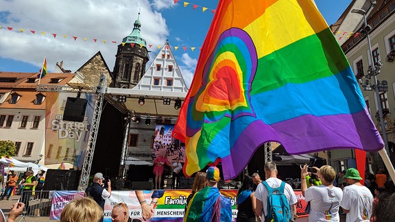 Eine Regenbogenfahne wird auf einem Marktplatz geschwenkt.  