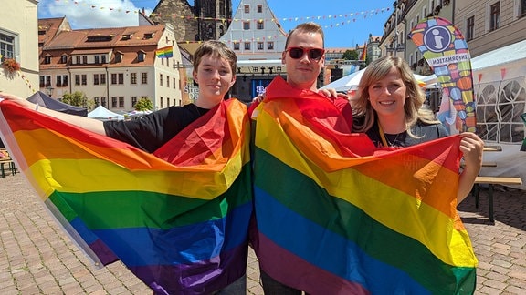 Zwei junge Männer und eine Frau halten Regenbogenfahnen und lächeln in die Kamera.  