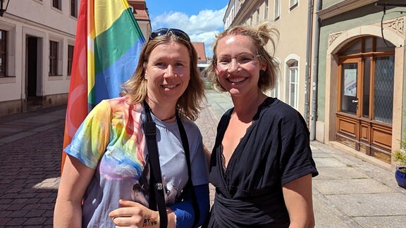 Zwei Frauen mit Regenbogenfahne lächeln in die Kamera. 