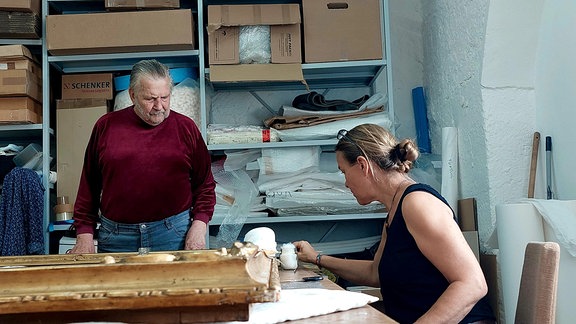 Ein Mann schaut einer Frau bei der Restaurierung eines Gemäldes zu.  