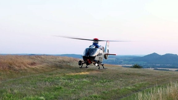 Ein Hubschrauber landet auf einem Feld in einer bergigen Landschaft.