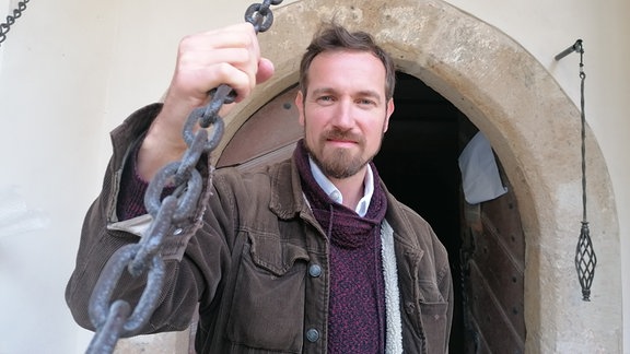 Gabor Schuster, ein Mann mit kurzen braunen Haaren und Bart mit lila Pullover und brauner Jacke hält eine eisene Kette in der Hand.