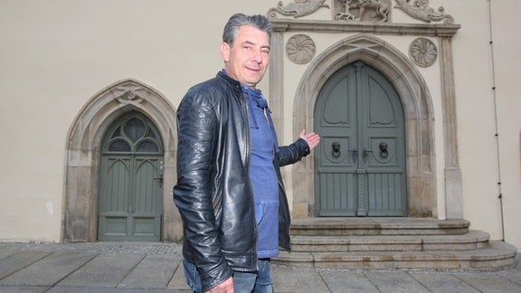 Ein Mann mit grauen Haaren und schwarzer Lederjacke steht vor einem historischen Hauseingang.