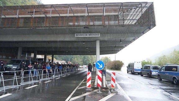 Unter der Überdachung des Grenzübergangs zwischen Schmilka in Sachsen und Hrensko in Tschechien haben sich Hunderte Menschen hinter einer Absperrung versammelt. Mehrere Polizeifahrzeuge und Beamte stehen daneben.