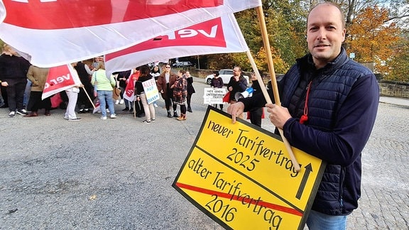 Ein Mann steht mit Verdifahnen und einem Plakat vor einer Klinik in der Mittagspause und protestiert gegen schlechte Arbeitsbedingungenund das Agieren ihrer Klinikleitung.