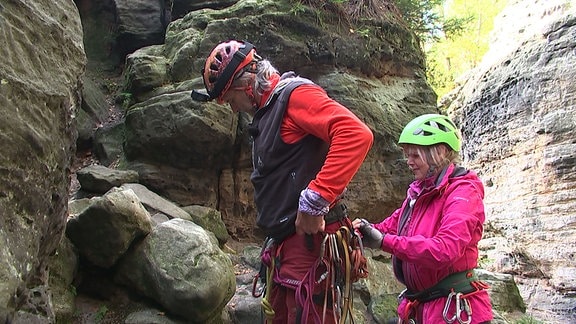 Eine Frau hilft einem Mann beim Anbringen von Kletterausrüstung. 