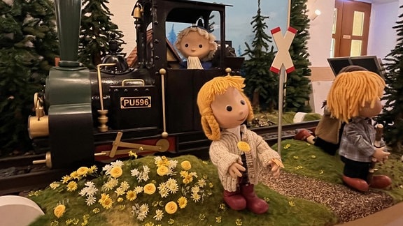 Sandmännchenfigur in einer Lokomotive und zwei Kinderfiguren, die Blumen pflücken