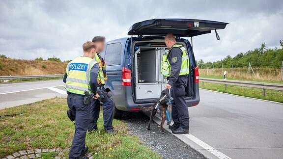 Drei Polizisten stehen mit einem Fährtenhund am geöffneten Heck eines Polizeitransporters. 