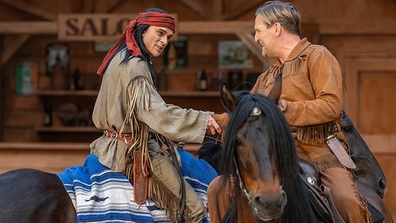 Ein Schauspieler mit langem, dunklem Haar in einem Kostüm, das der Kleidung amerikanische Ureinwohner nachempfunden ist, sitzt auf einem Pferd und reicht einem blonden Schauspieler in Cowboyjacke, der ebenfalls auf einem Pferd sitzt, die Hand.