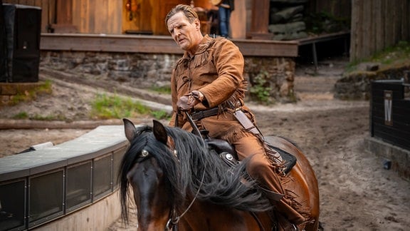 Ein blonder Schauspieler in Cowboykostüm reitet auf einem Pferd.