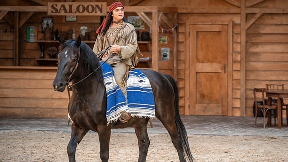 Ein Schauspieler mit langem, dunklem Haar in einem Kostüm, das der Kleidung amerikanische Ureinwohner nachempfunden ist, reitet auf einem Pferd.