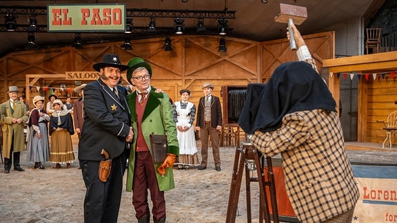 Ein Schauspieler in Sheriff-Kostüm und ein Schauspieler mit grünem Mantel und Zylinder posieren vor einer historischen Kamera.