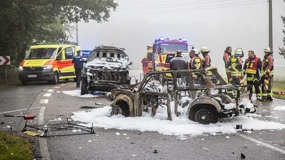 Ein ausgebrannter Trabi steht auf einer Straße. Um den Wagen stehen Feuerwehrleute. Im Hintergrund sind Einsatzfahrzeuge zu sehen sowie ein größeres Fahrzeug, ebenfalls mit Brandschäden.  