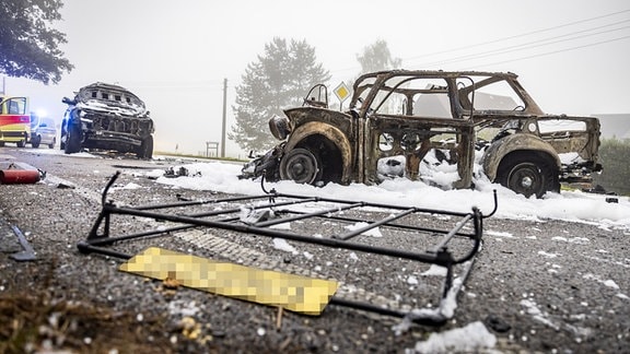 Ein ausgebrannter Trabi steht auf einer Straße. Im Hintergrund ist ein gößeres Auto zu sehen, ebenfalls mit Brandschäden. Im Vordergrund liegt ein Blechschild auf dem "Schorschi" steht