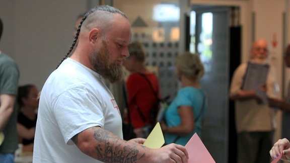 Ein Mann mit tätowierten Unterarmen steht an einer Wahlurne und will seine Wahlzettel einwerfen.  