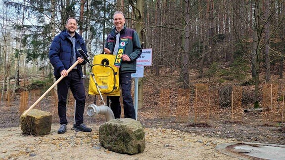 Thomas Kunack, Bürgermeister von Bad Schandau (links)  und Uwe Borrmeister, Leiter der Nationalpark- und Forstverwaltung Sächsische Schweiz, stehen an der Wasserentnahmestelle einer Löschwasserzisterne im Wald und halten eine Waldbrandhacke und einen Löschwasserrucksack in den Händen.