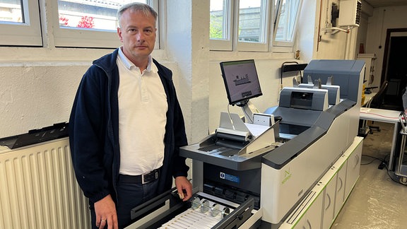 Ein Mann mit grauen Haaren und weißem Poloshir steht vor einer länglichen Druckmaschine.