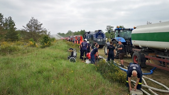 In einer Heidelandschaft stehen hintereinander Feuerwehrlöschfahrzeuge, ein Wasserwerfer der Polizei und ein Traktor mit Wassertank-Anhänger. Im Hintergrund versprühen Kreisregner Wasser.