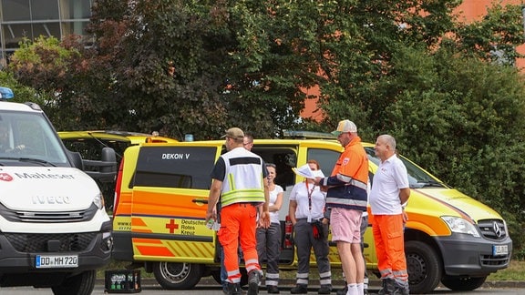 Feuerwehrautos stehen in einer langen Schlange vor einem Betriebsgelände. Rettungskräfte stehen vor den Autos und schauen angespannt.