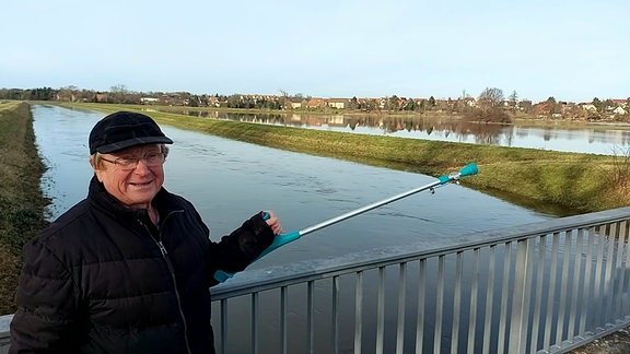 Auf einer Brücke steht ein Rentner und zeigt mit seiner Gehilfe (Krückstock) in der linken Hand auf eine überschwemmte Auenlandschaft. 