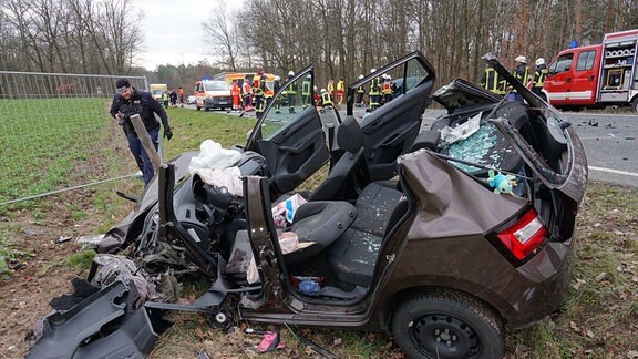 Eine Person nähert sich einem total zerstörten Autowrack. Im Hintergrund Rettungswagen und Personal