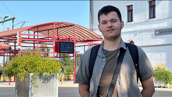 Ein junger Mann steht an einem Bahnhof und schaut in die Kamera.