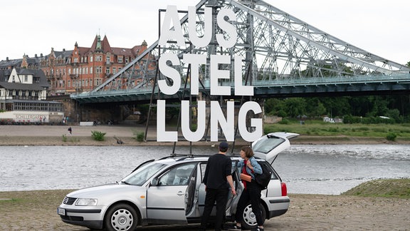 Ina Weise (r) und Christian Göthner, Kuratoren, stehen unter dem Schriftzug "Ausstellung" vor einem VW Passat (Baujahr 1997) am Ufer der Elbe vor der Elbbrücke Blaues Wunder. 