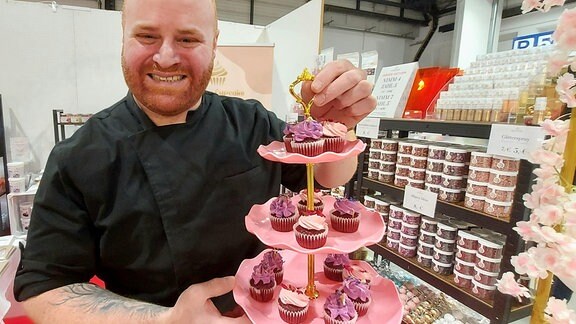 Ein Mann mit rötlichem, kurzen Vollbart blickt in die Kamera. In der linken Hand hält er eine Etagere. Die ist voller leckerer rosafarbener Cupcakes.