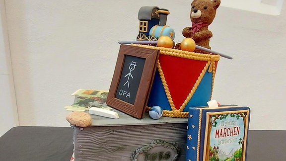 Eine Szenerie nimmt Bezug auf deutsche Märchen. Auf einer Holzkiste sitzt ein Teddy, ein Märchenbuch lehnt an der Kiste und eine Schiefertafel trägt dass Wort Opa. Das Ganze ist komplett aus Kuchen, Schokolade ud Marzipan.