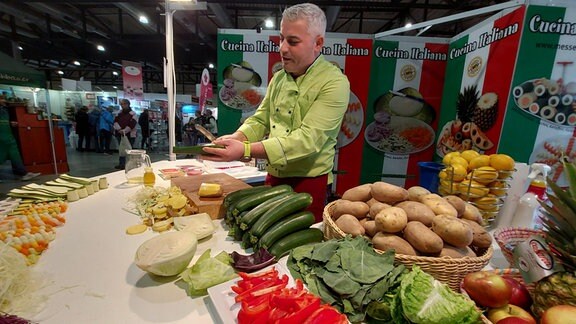 Ein Mann aus Italien steht an einem Messestand vor viel Gemüse und Obst und zeigt mit einem speziellen Schäler Schältechniken.