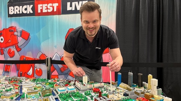 Ein junger Mann mit Bart steht vor einer Miniaturstadt aus Lego und lächelt in die Kamera. 