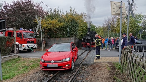 PKW auf Gleisen der Lößnitzgrundbahn, dahinter Einsatzkräfte, Feuerwehr, Dampflok und Schaulustige