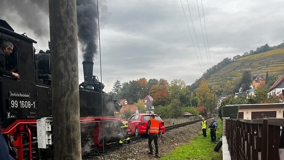 Dampflok vor PKW auf Gleisen der Lößnitzgrundbahn, daneben Einsatzkräfte