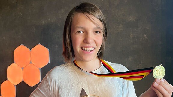 Ein Junge mit Zahnspange und längeren Haaren zeigt seine Goldmedaille in die Kamera.