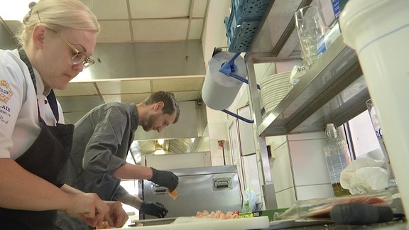 In einer Restaurant-Küche arbeiten ein Koch und eine Köchin mit Fisch und Salat.