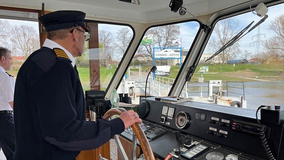 Ein Mann mit Kapitänskleidung steht am Steuerrad eines Schiffes